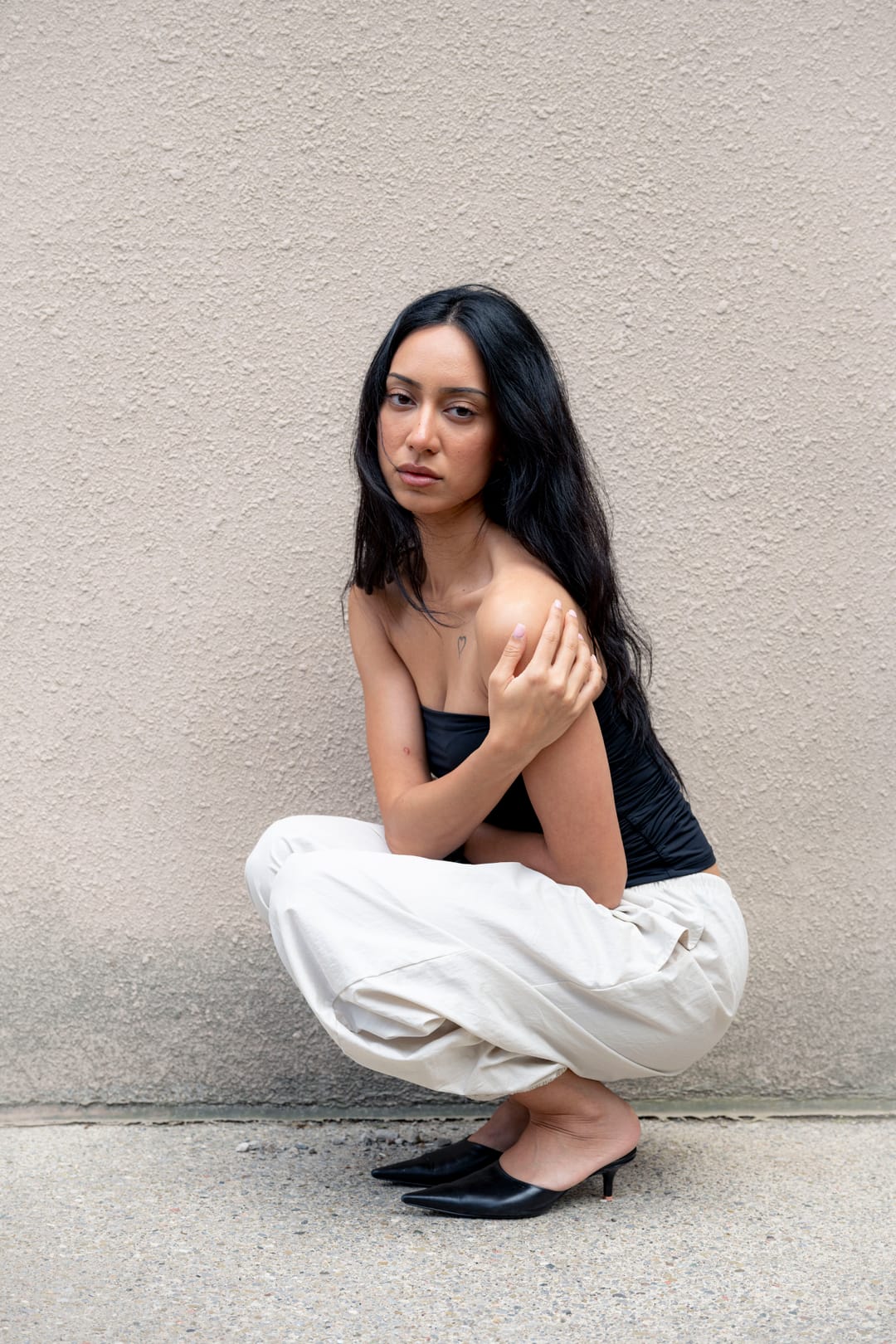Model in a black top and cream trousers crouching against a pale stucco wall