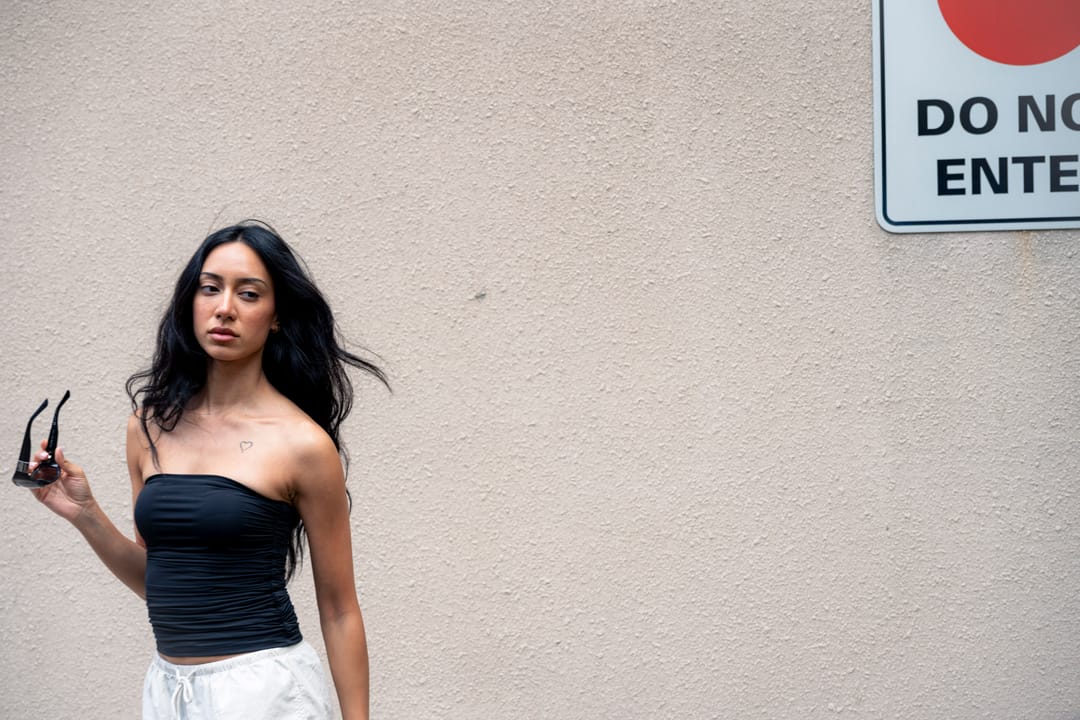 Model holding sunglasses in front of a pale wall beside a do-not-enter sign