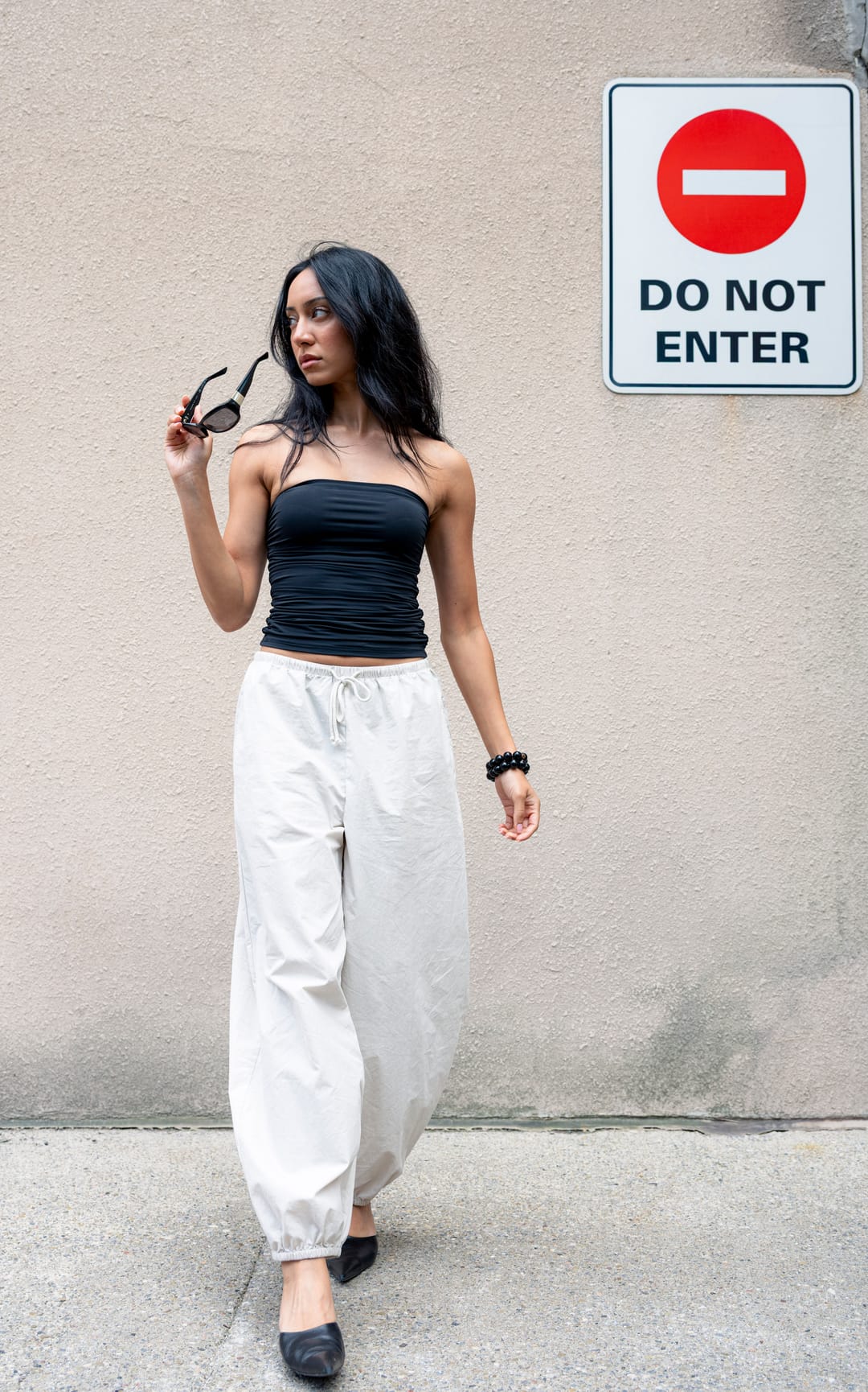 Model in a black top and cream trousers lifting sunglasses in front of a do-not-enter sign