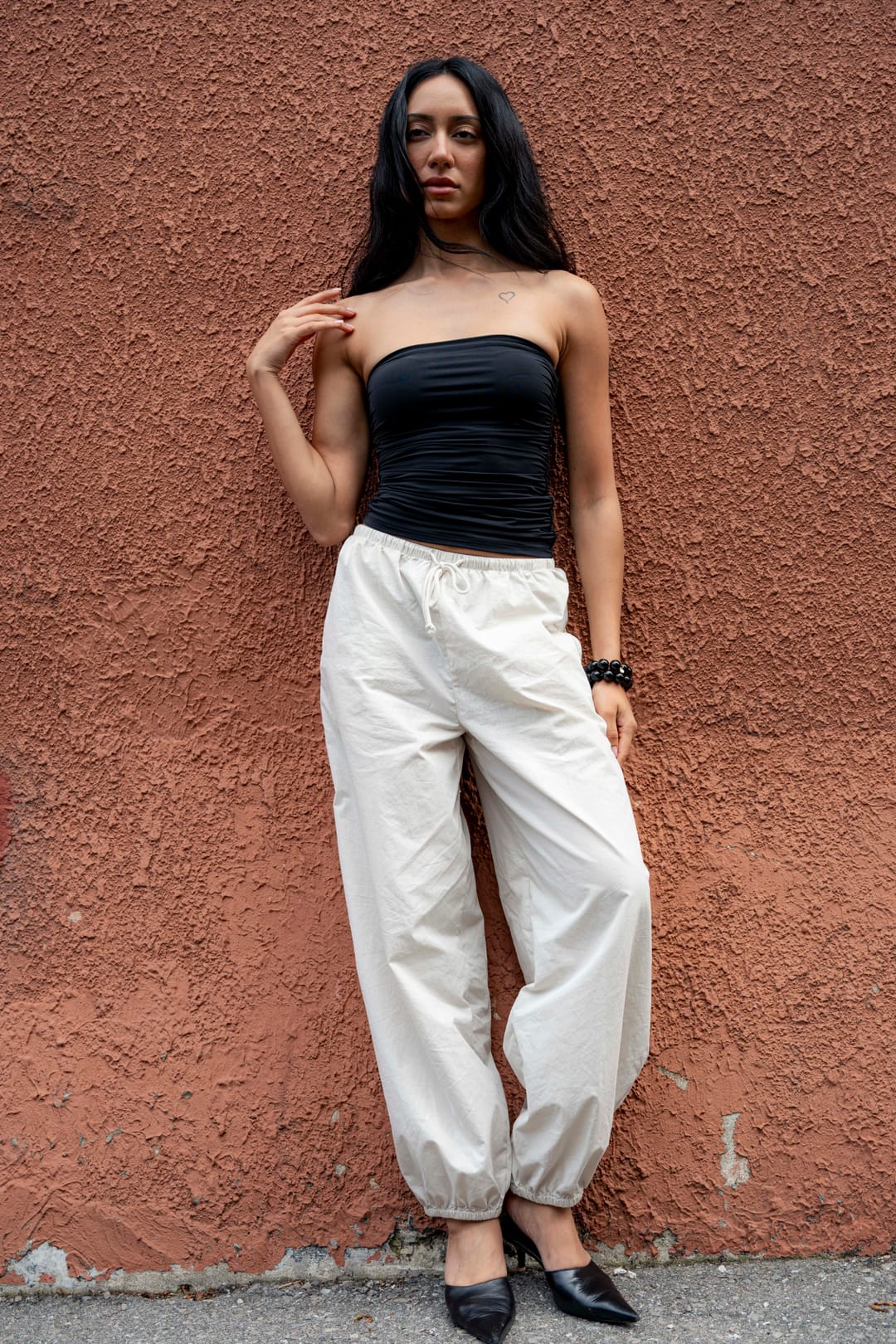 Model in a black top and cream trousers leaning against a terracotta wall