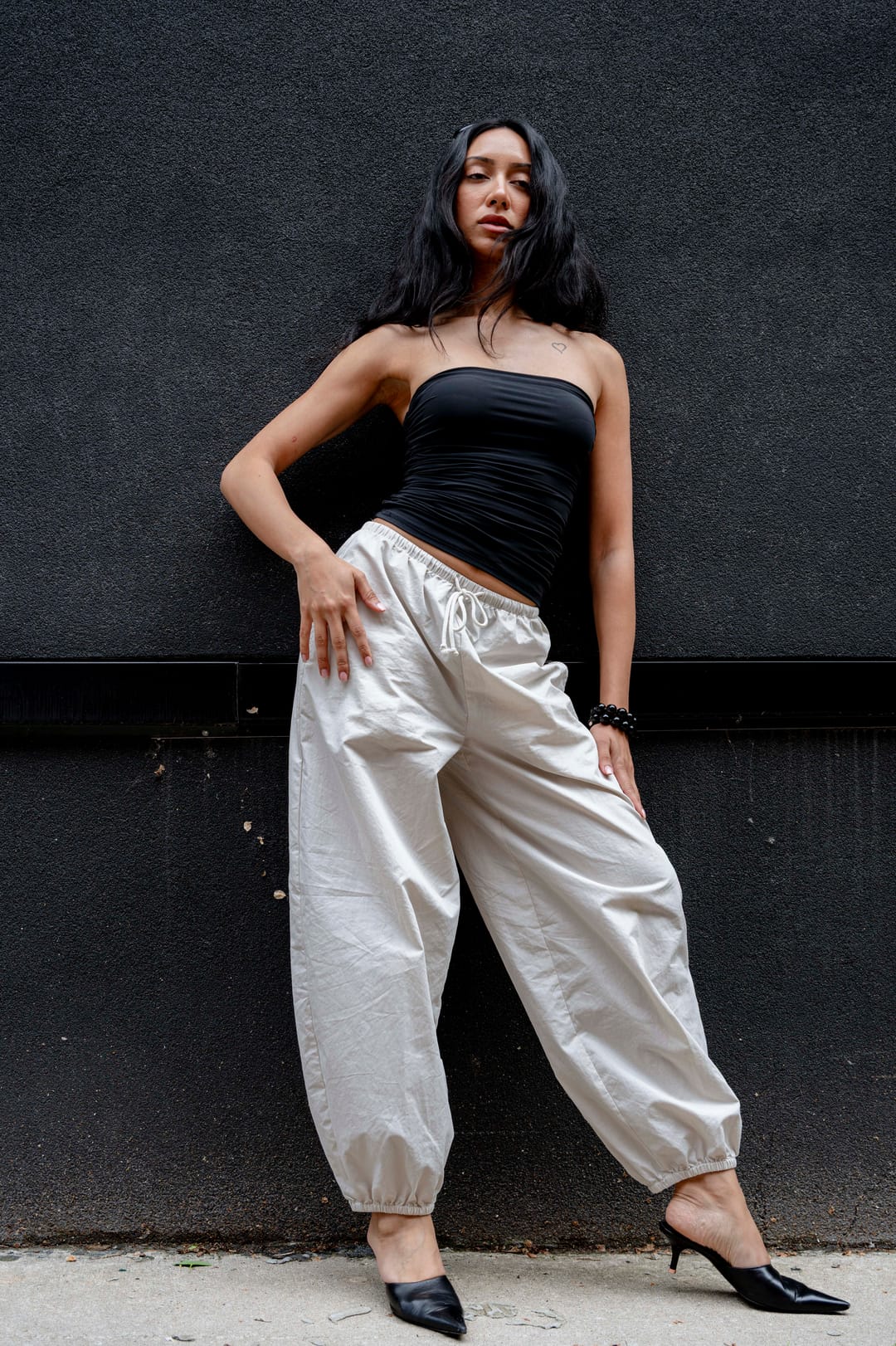 Model leaning on a black wall in cream trousers and black heels