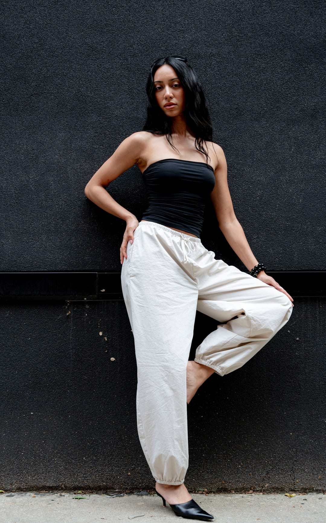 Model in a black top and cream trousers posed with one knee bent against a dark wall