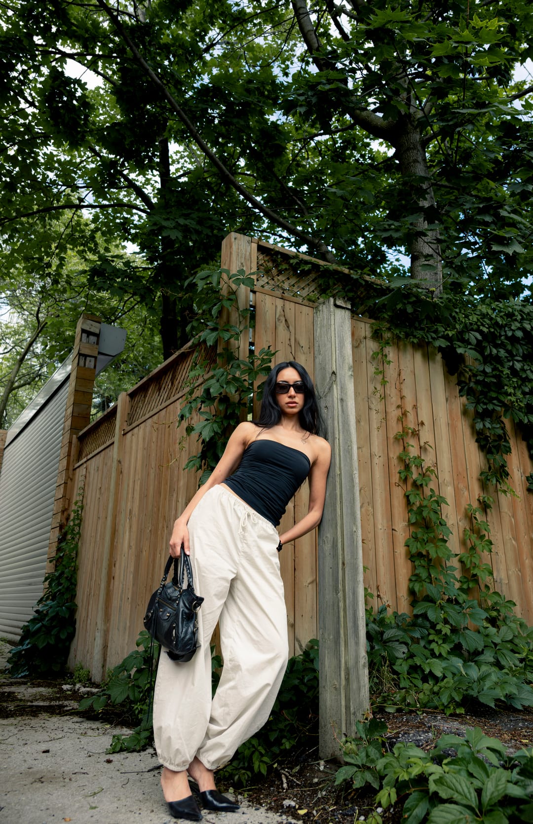 Model with a black handbag leaning on a wooden fence beside garden greenery