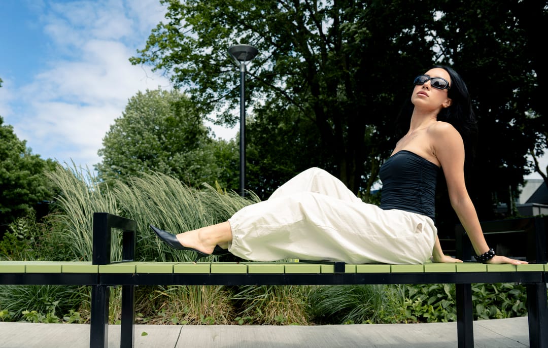 Model reclining on a park bench beneath a blue sky, trees behind