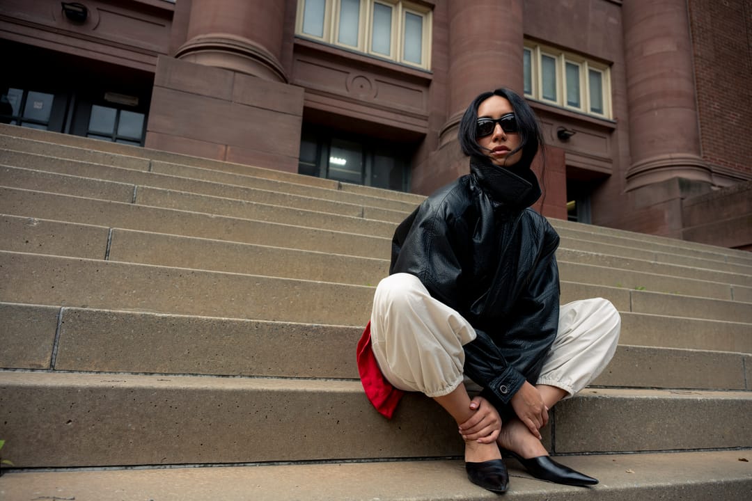 Model in a black leather jacket seated on stone steps in front of a red brick building