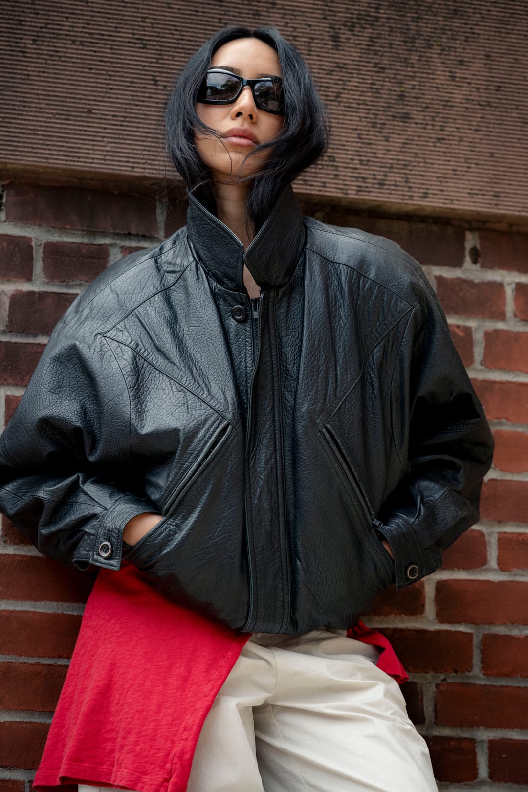Model in a black leather jacket with a red shirt tied at the waist, leaning on a brick wall