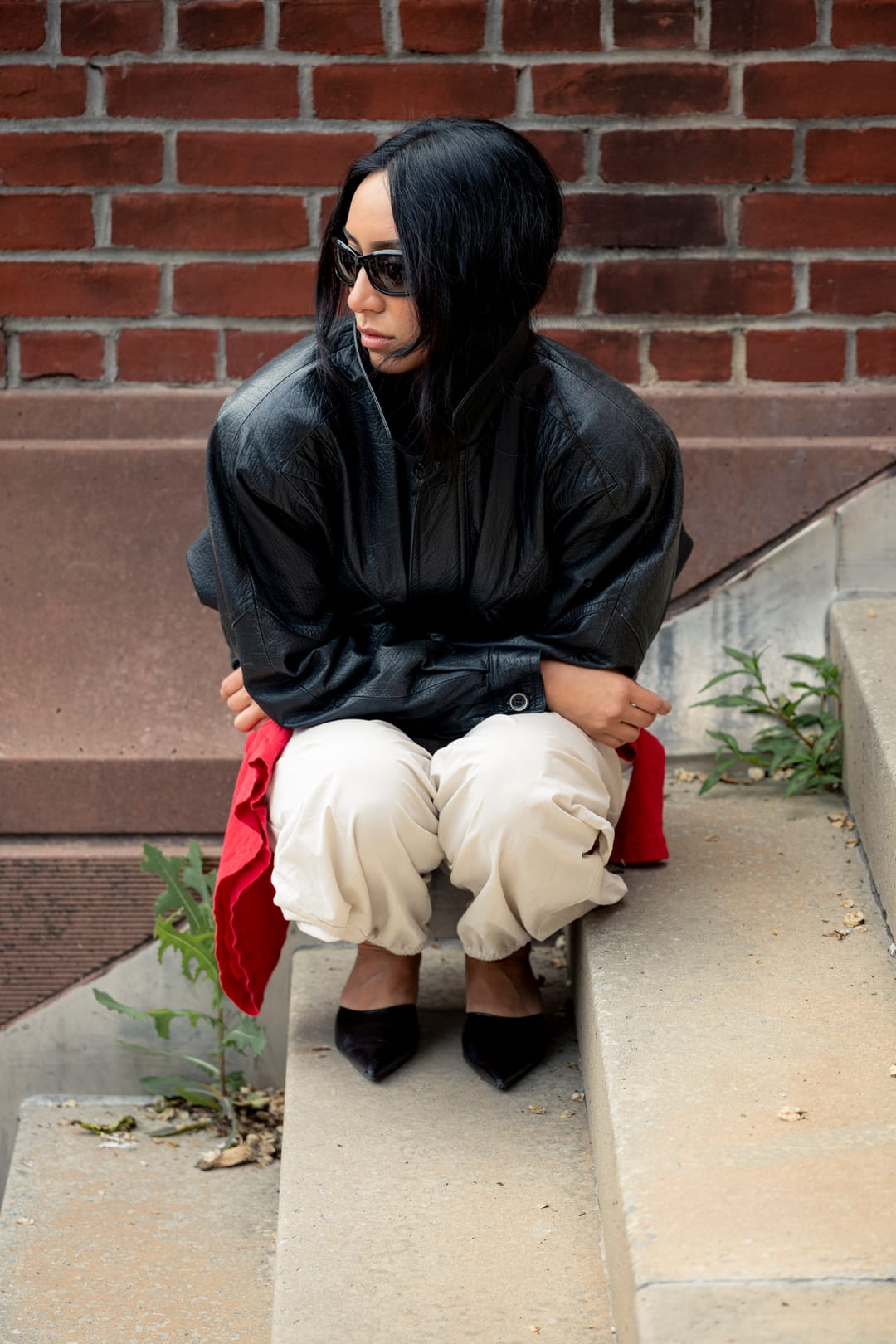 Model crouching on stone steps beside a brick wall in a leather jacket and sunglasses