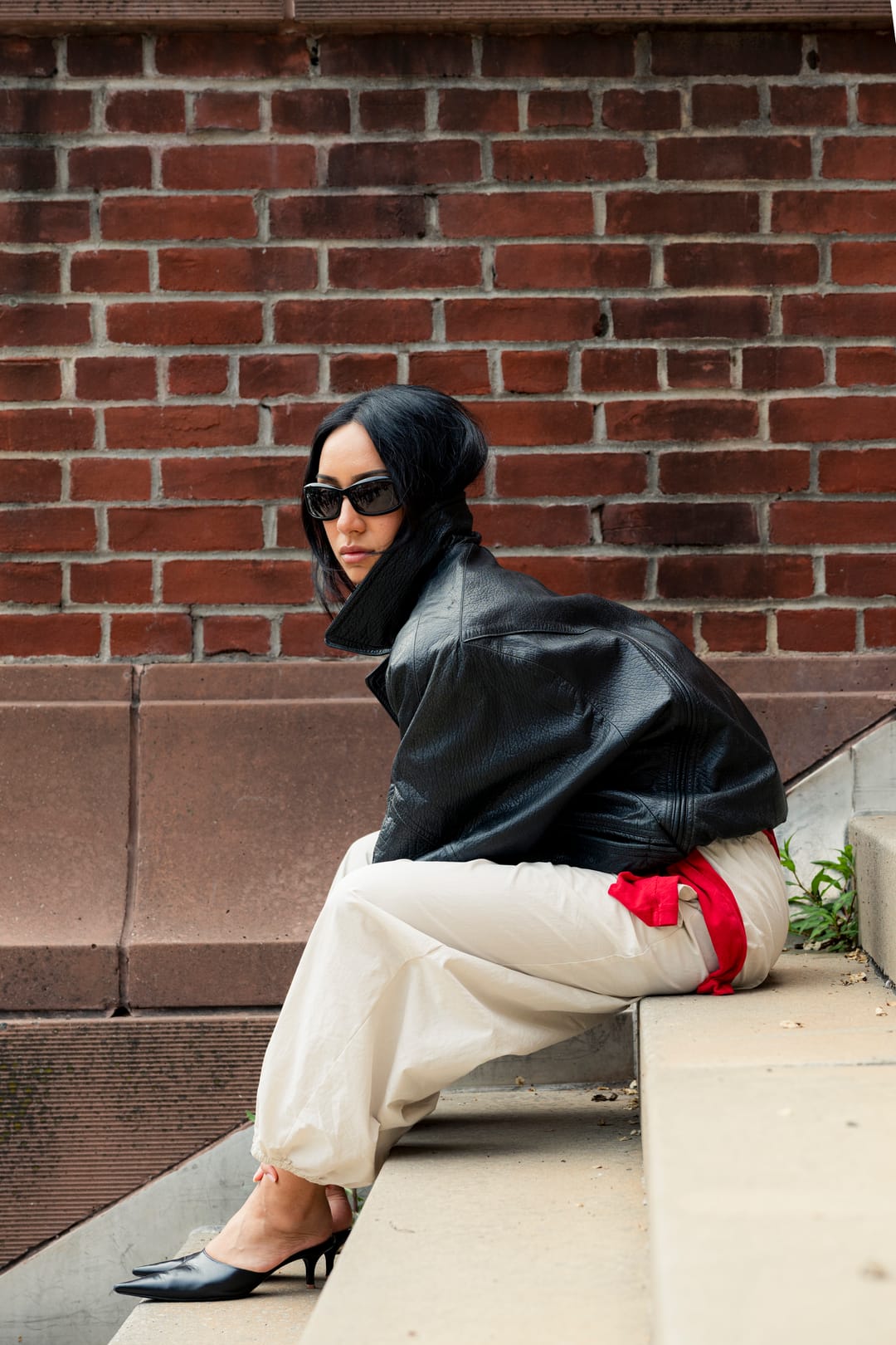 Model seated on brick steps in a leather jacket, looking back over a shoulder