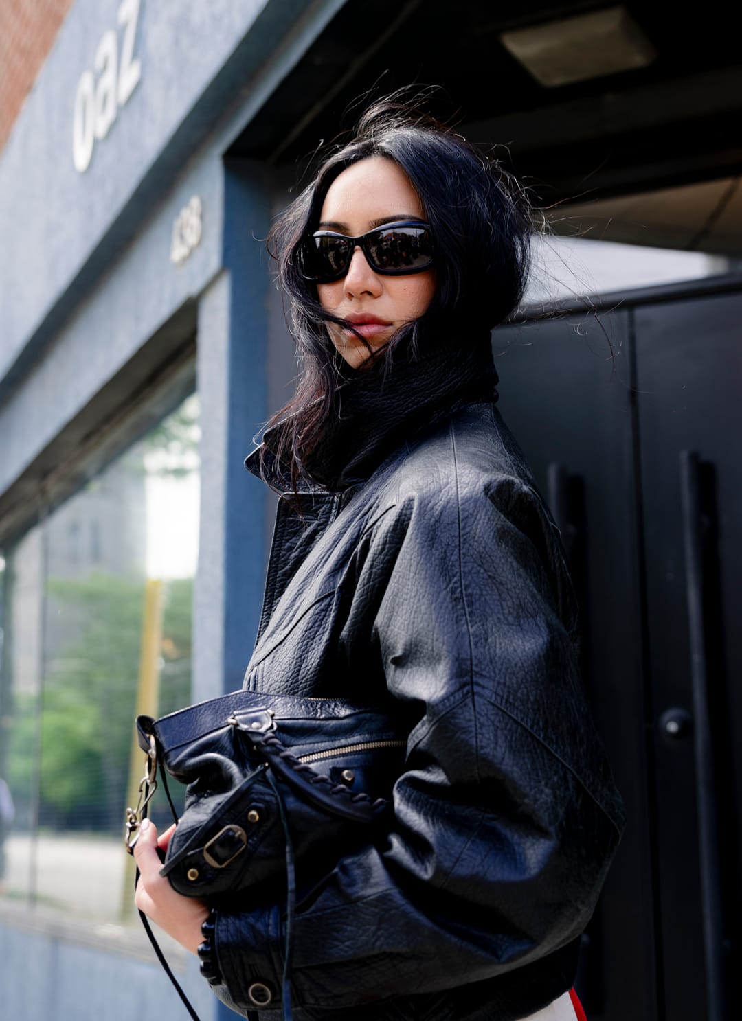 Model in a leather jacket holding a black bag in front of a blue storefront