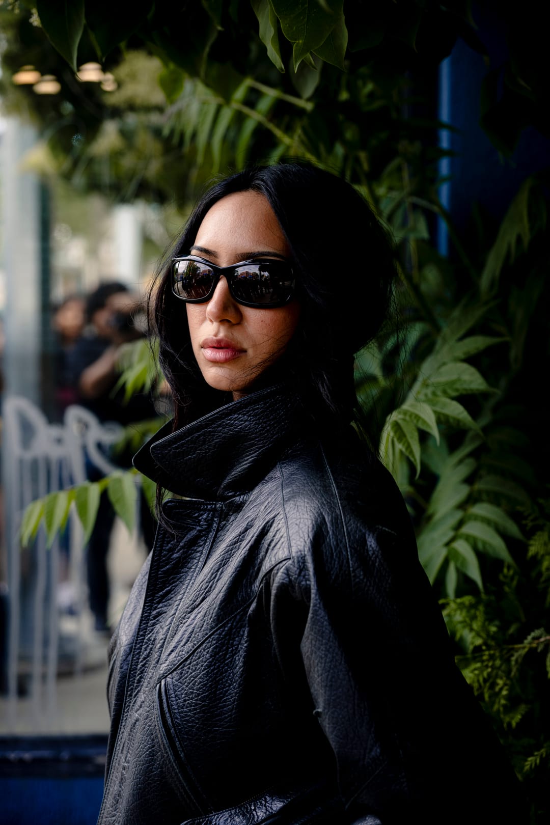 Close portrait of a model in sunglasses and a high-collared leather jacket beside green ferns