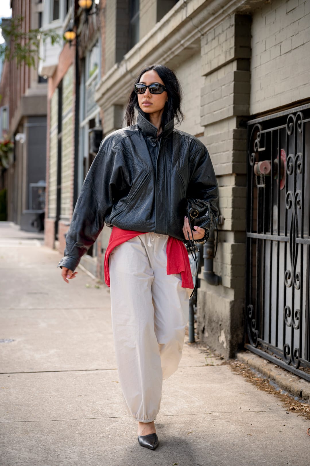 Model in a leather jacket and cream trousers walking down a sidewalk beside brick buildings