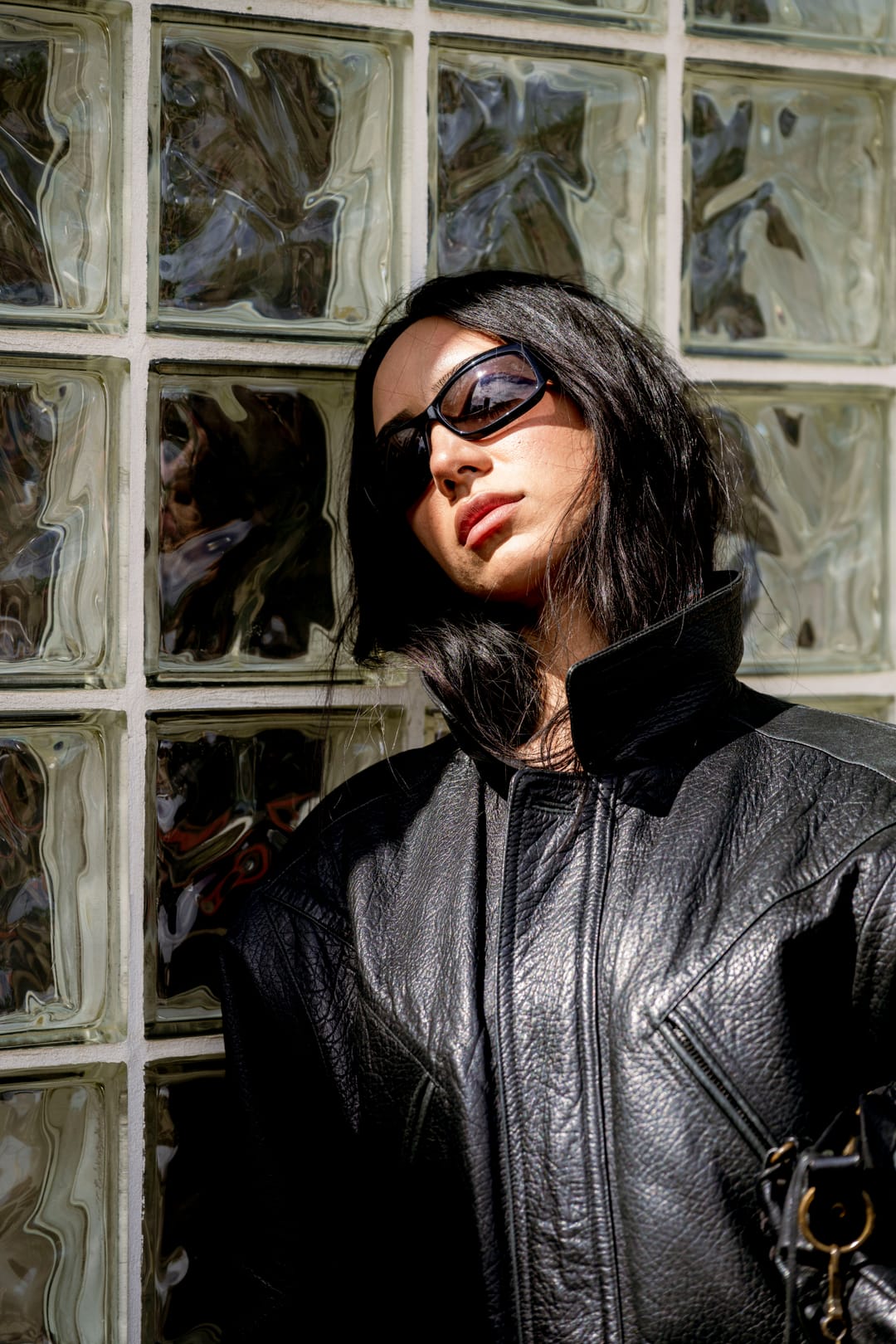 Model in sunglasses and a leather jacket leaning against a glass-block window