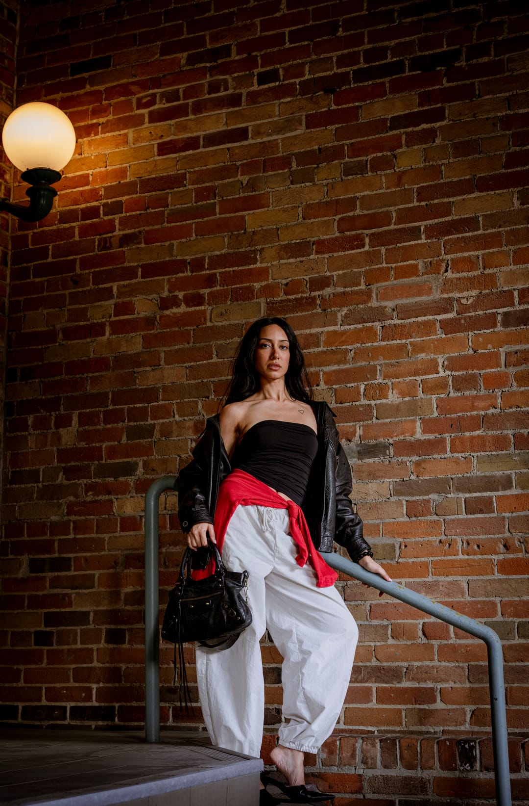 Model in a leather jacket standing on brick steps beneath a glowing wall lamp