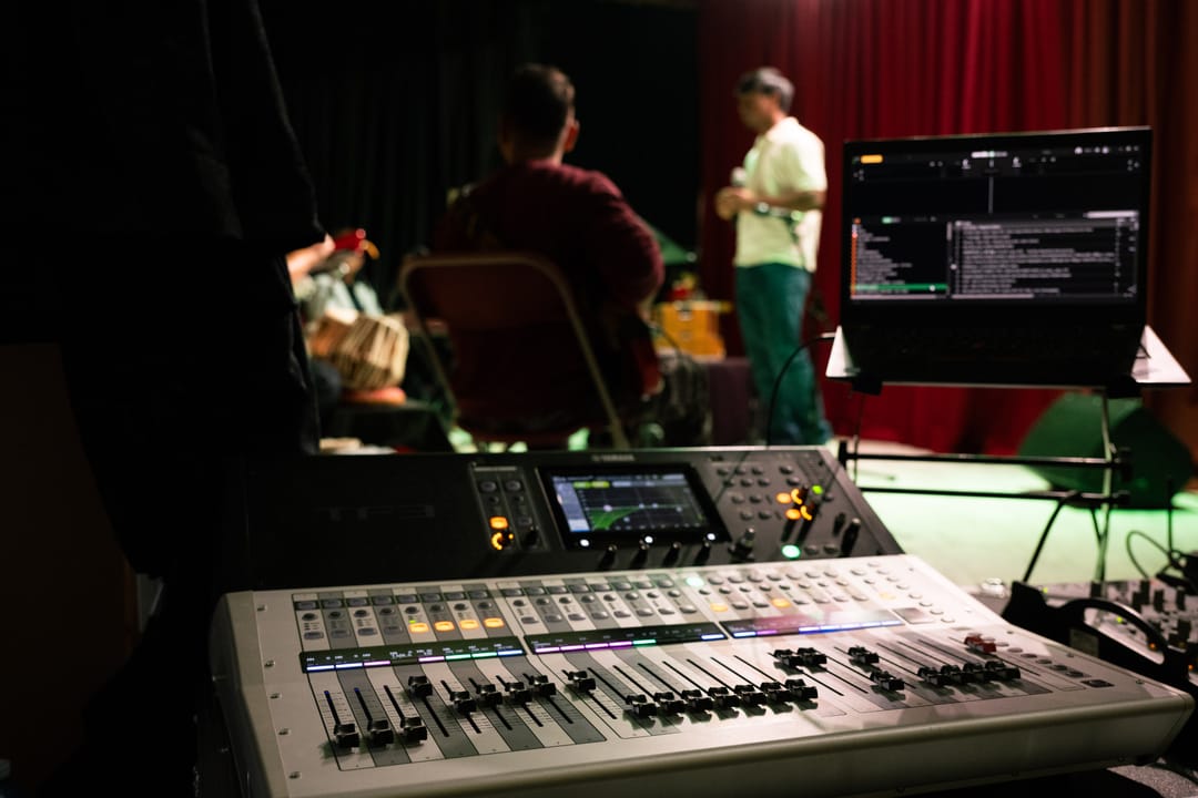 A sound mixing console at the side of a stage, a technician in the background