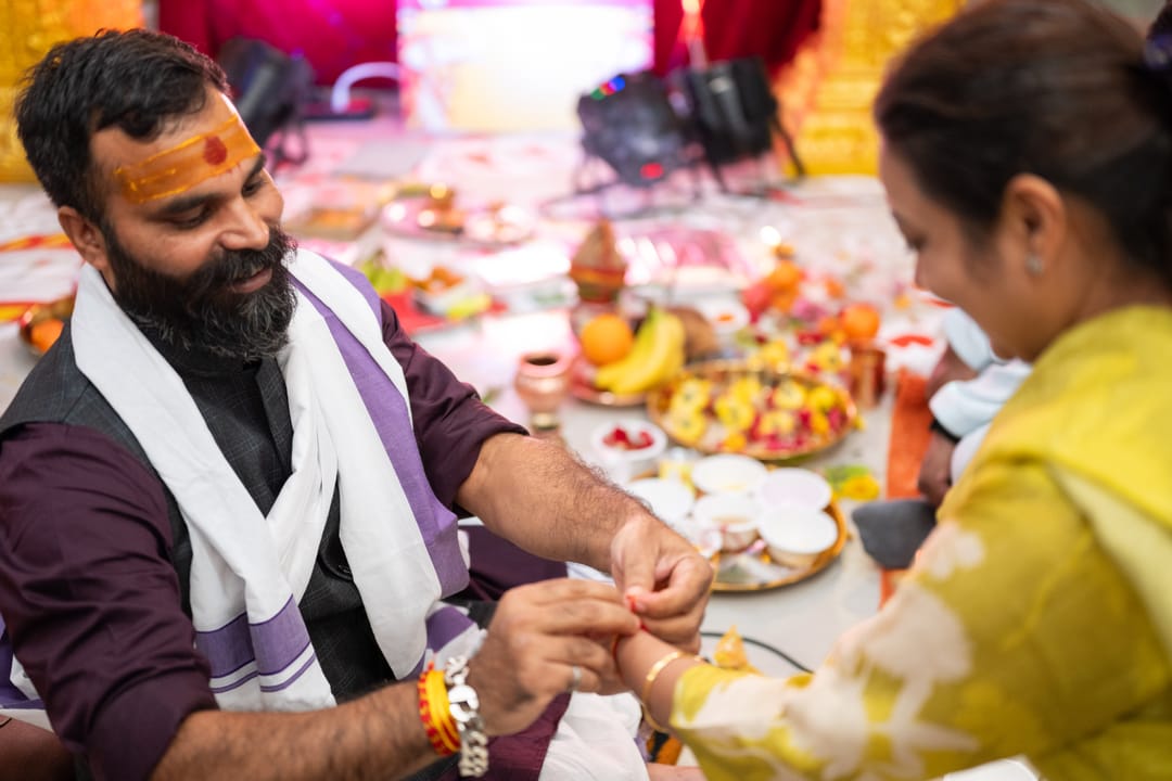 A man in a white stole tying a thread on a woman’s wrist above a table of fruit