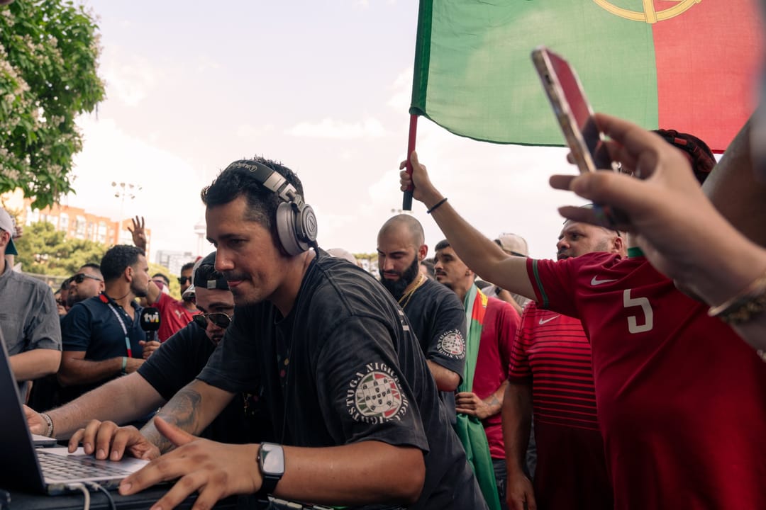 A DJ in headphones plays at a laptop surrounded by Portugal fans in red jerseys, one waving a flag and another filming on a phone