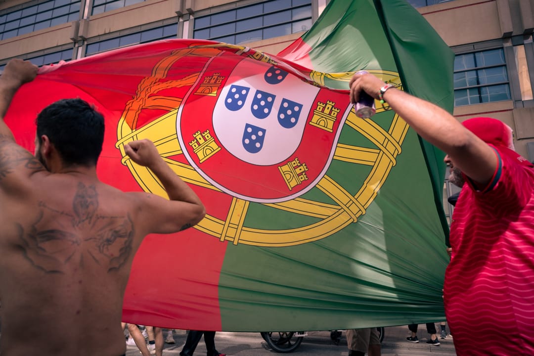 Two supporters hold up an enormous Portugal flag on a city street, one of them shirtless with a tattooed back