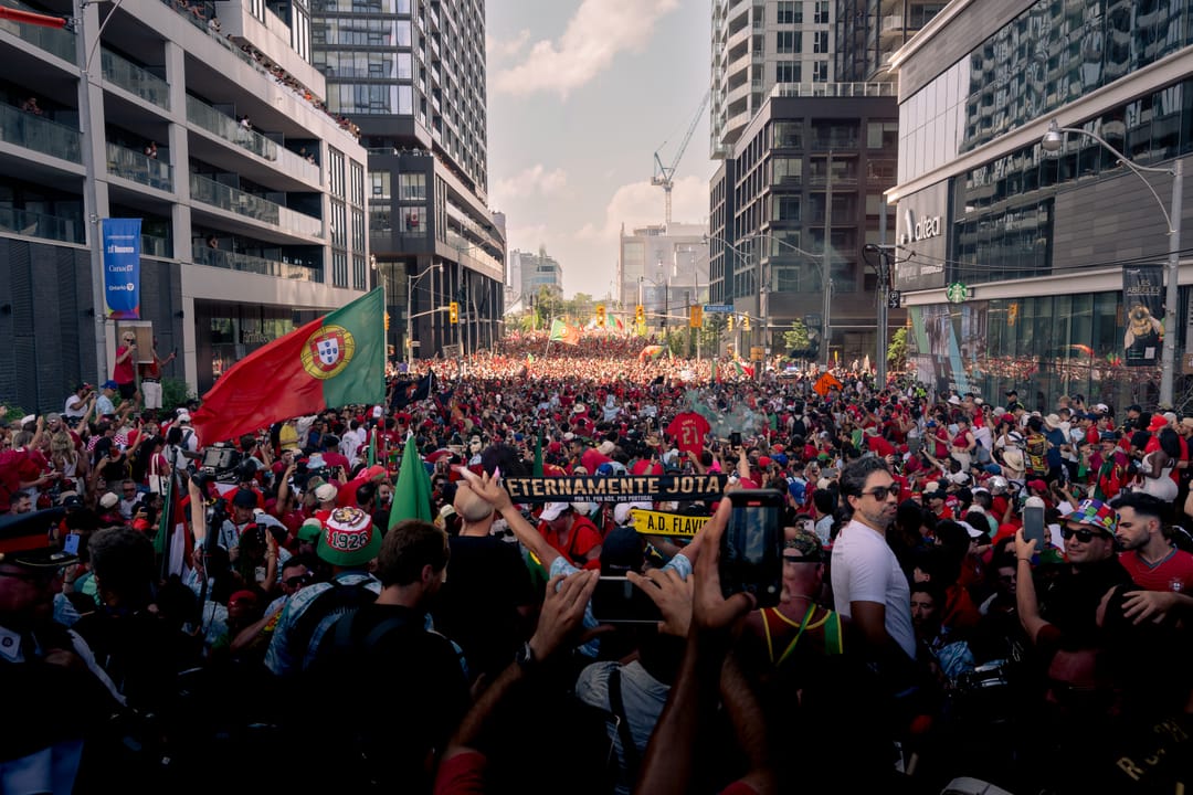 A packed street of fans marching between apartment towers, a Portugal flag in the foreground