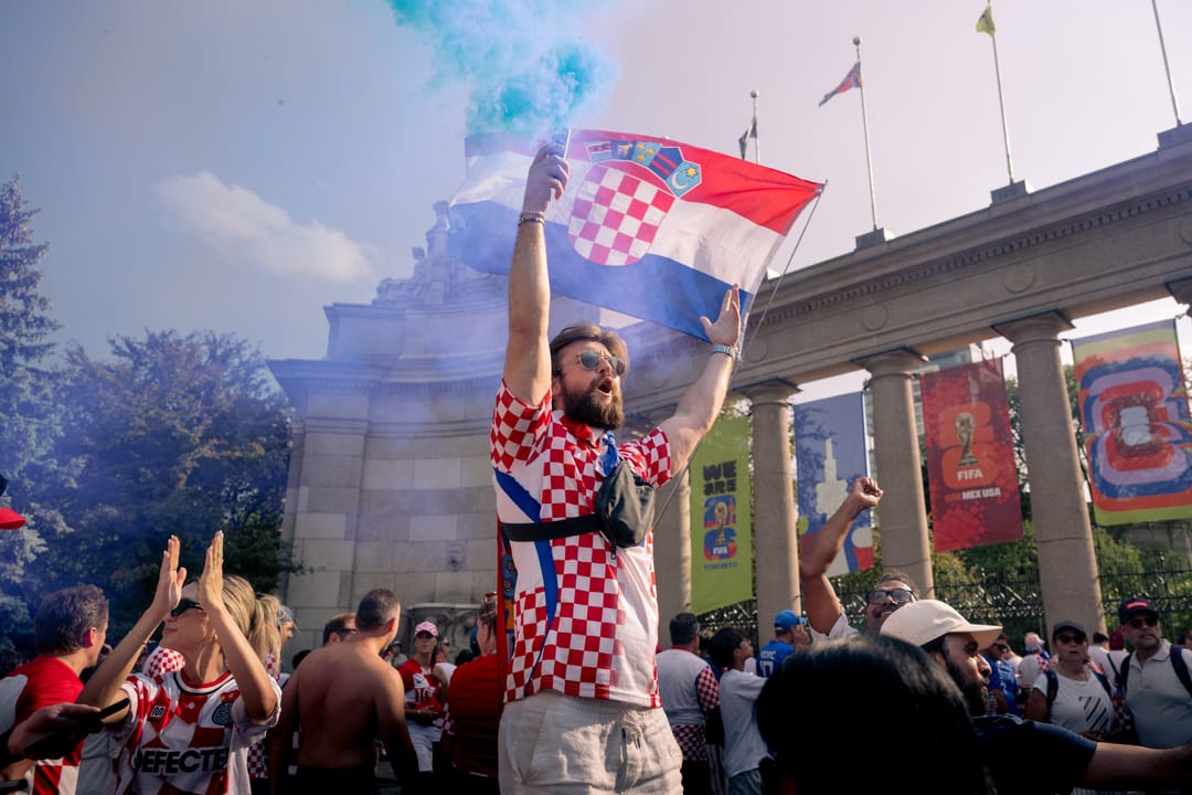 A fan in a Croatia jersey holding a flag and a blue smoke flare in front of a stone colonnade