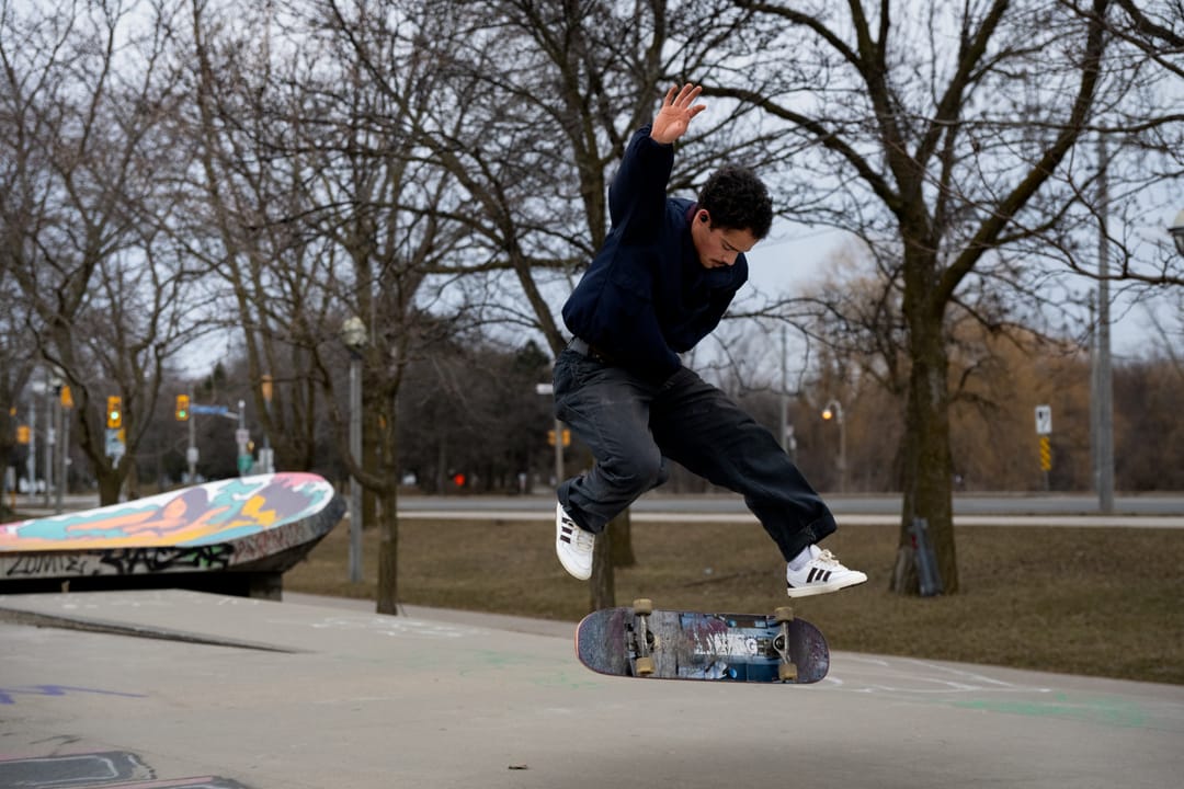 A skateboarder flipping a board past bare trees near a painted bowl