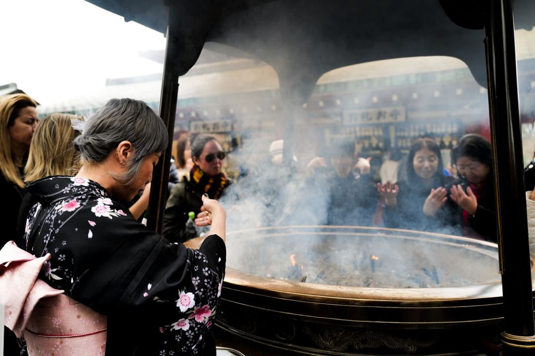 A woman in a black floral kimono with a pink sash reaches toward a large smoking incense burner as other visitors wave the smoke toward themselves