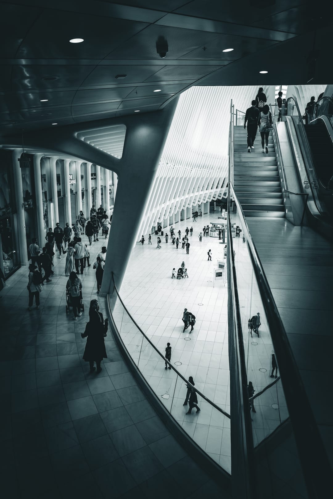A curved white transit hall seen from above, people crossing the floor and escalators