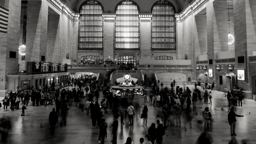 Black and white view of a vast station concourse, crowds blurred by a long exposure around a central information booth with a clock